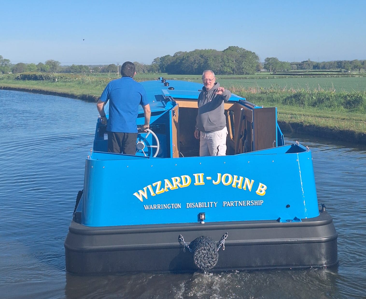 two men on the blue, wide-beam narrow boat The Wizard II - John B on the river offering accessible rides to everyone.